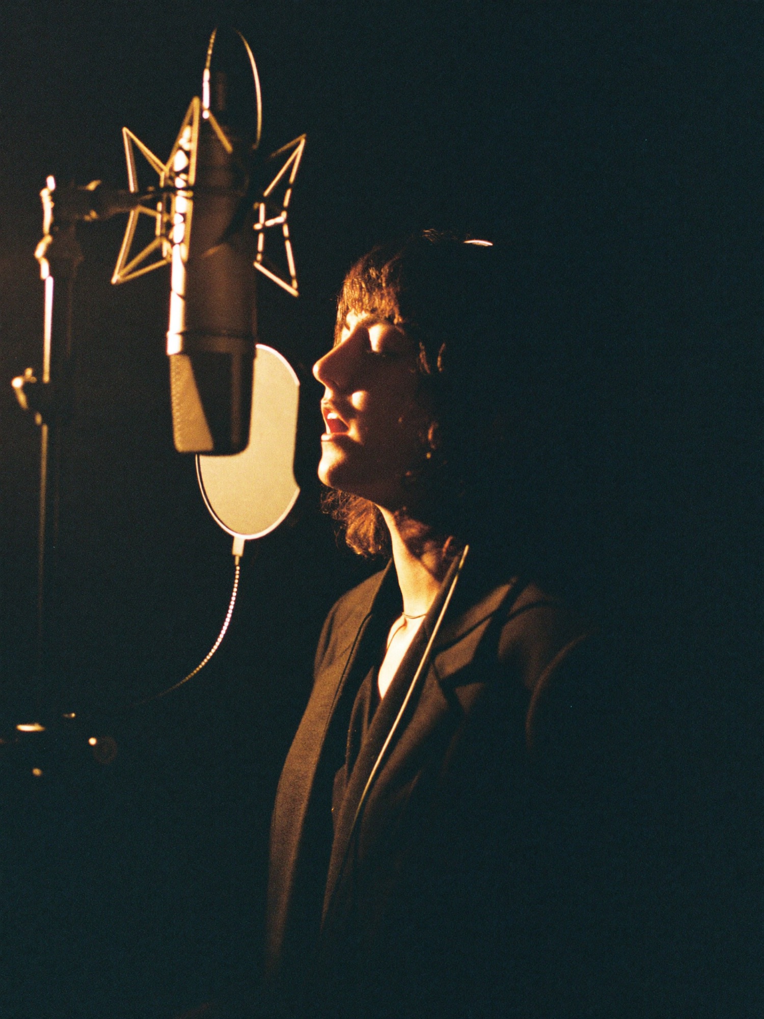 A vocalist recording into a condenser microphone in a dim booth with warm rim light
